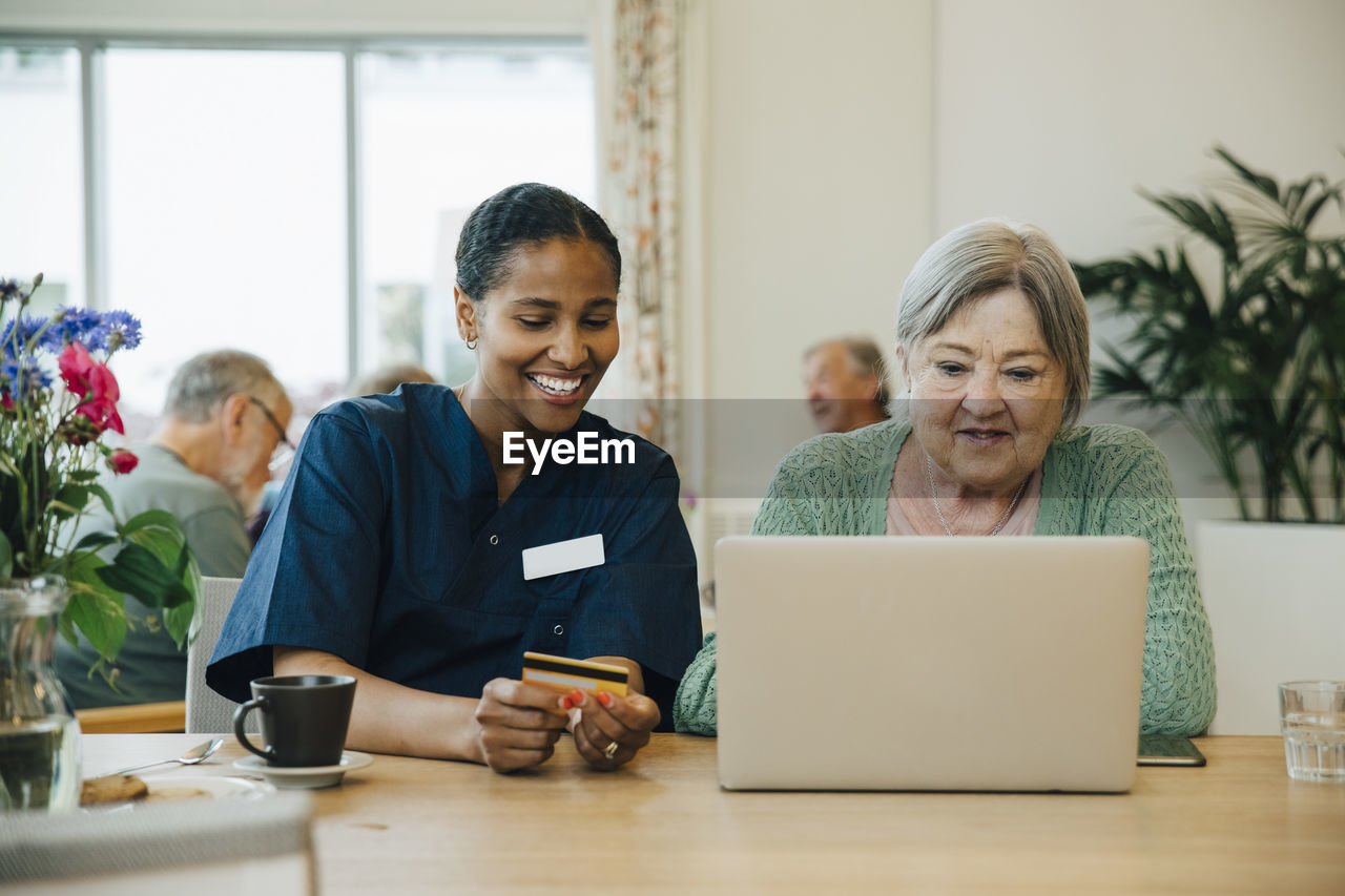 Smiling female caregiver assisting senior woman doing online shopping with credit card and laptop in nursing home