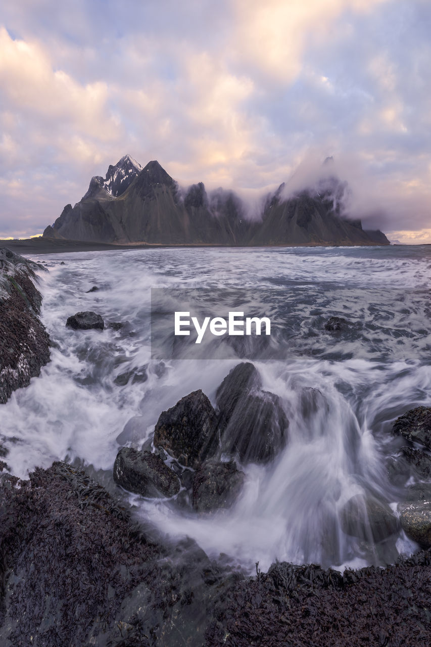 Picturesque scenery of waving sea washing rough shore near high snowy mountains reaching cloudy sky