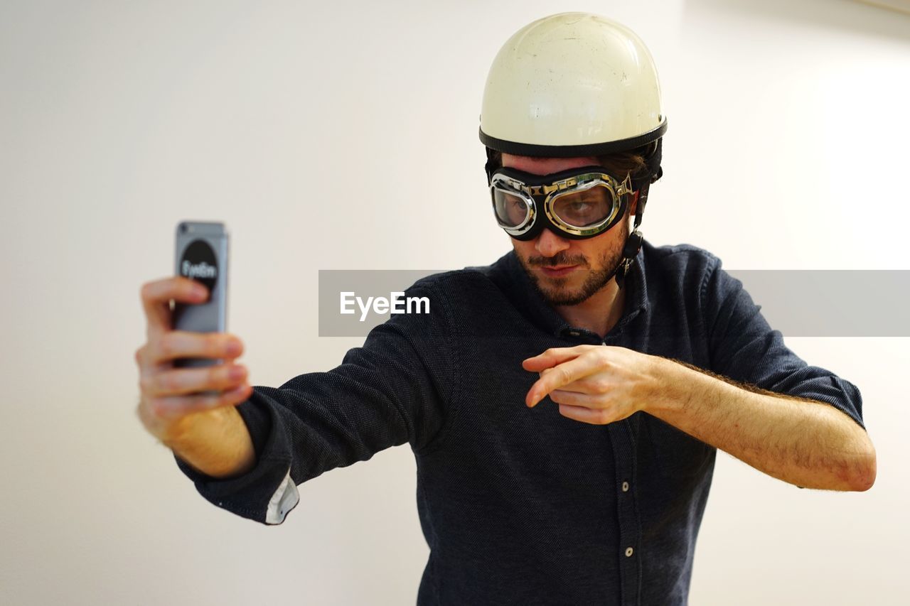 Young man in aviator glasses and helmet taking selfie against white wall