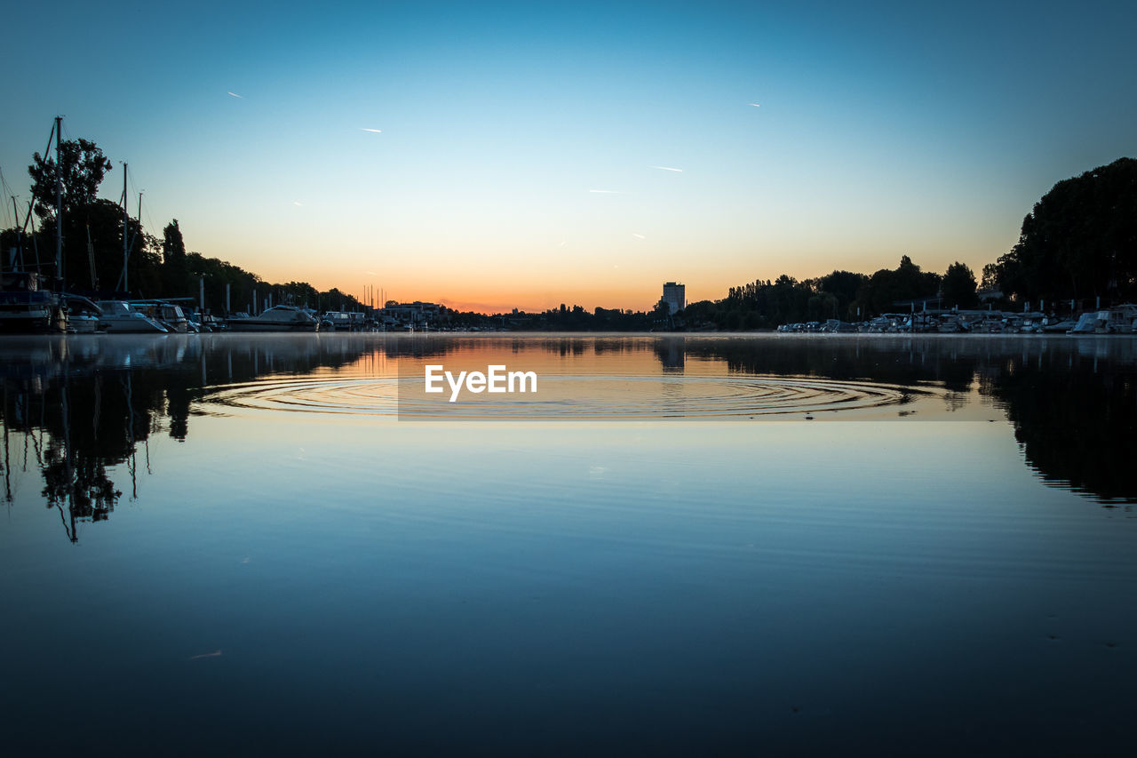 REFLECTION OF SILHOUETTE TREES IN LAKE AGAINST SUNSET SKY