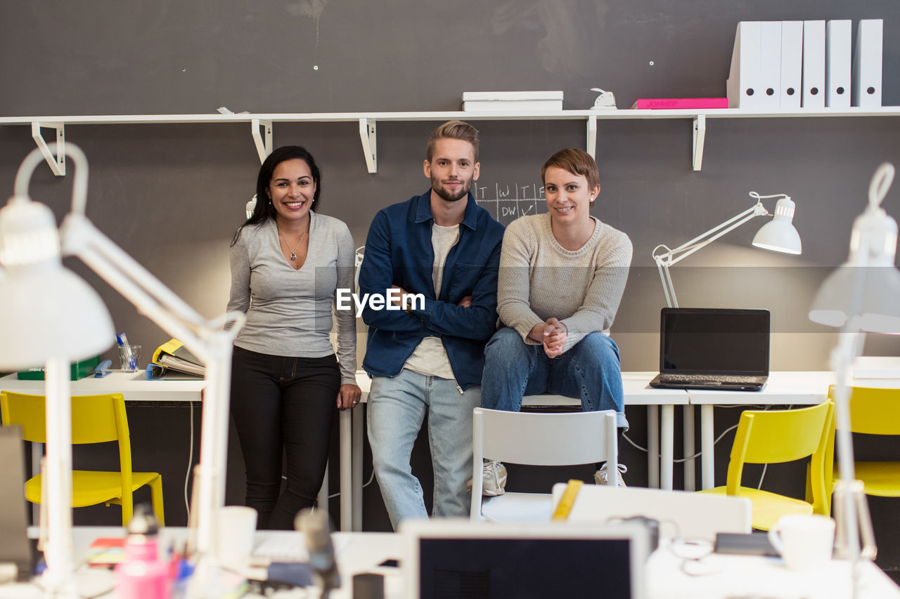 Portrait of confident multi-ethnic business colleagues sitting on desk against wall at creative office