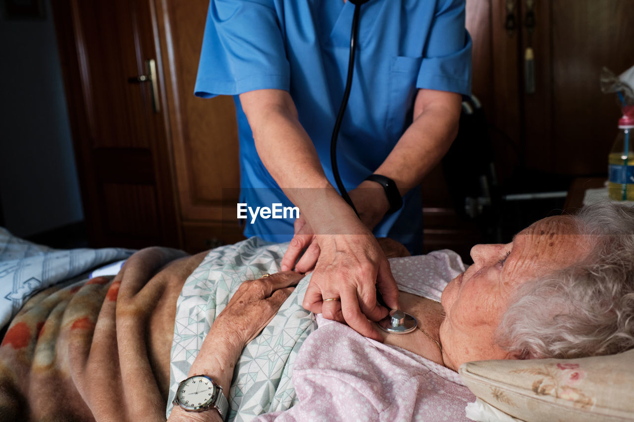 Crop unrecognizable nurse in uniform measuring heart rate of elderly female patient with special stethoscope lying on bed in bedroom