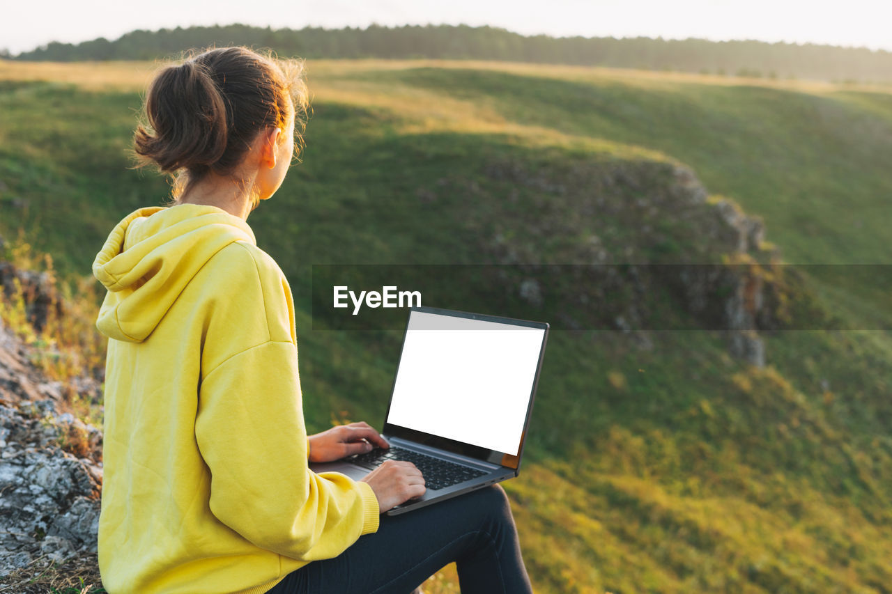 Crop photo of young woman traveler freelancer in yellow hoodie with white screen opened laptop 