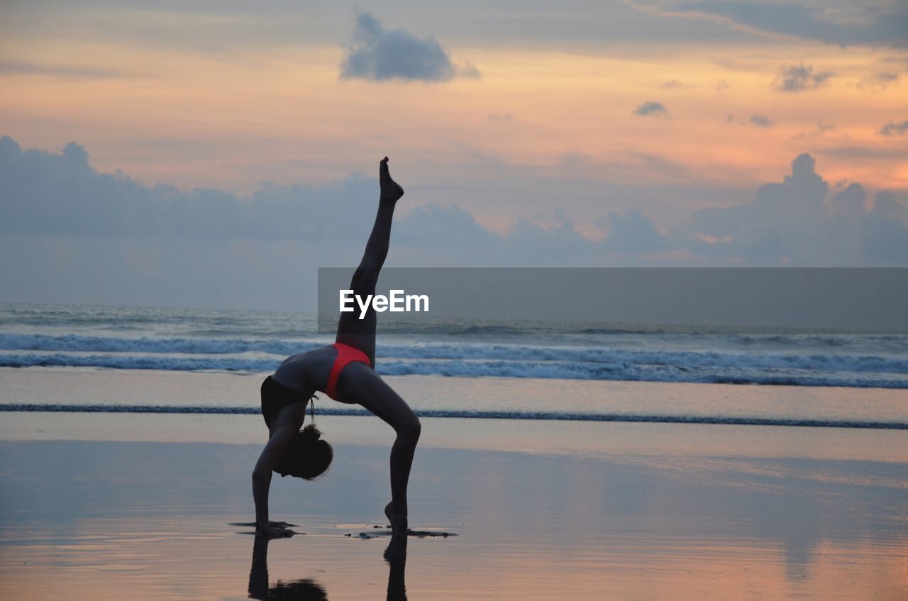 Full length of woman exercising at beach against sky during sunset