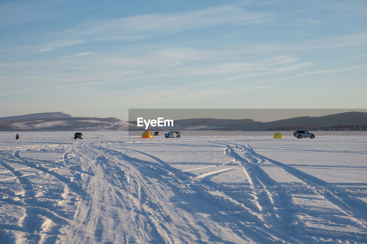 Trace from cars on a snowy big lake with fishermen in the background.