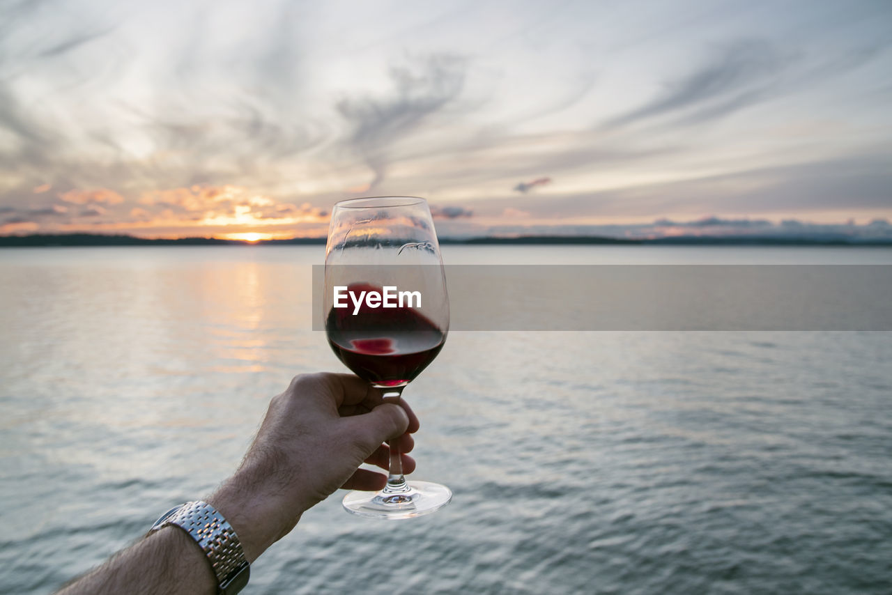 Point of view hand swirling red wine under a swirling cloud sunset over sea.