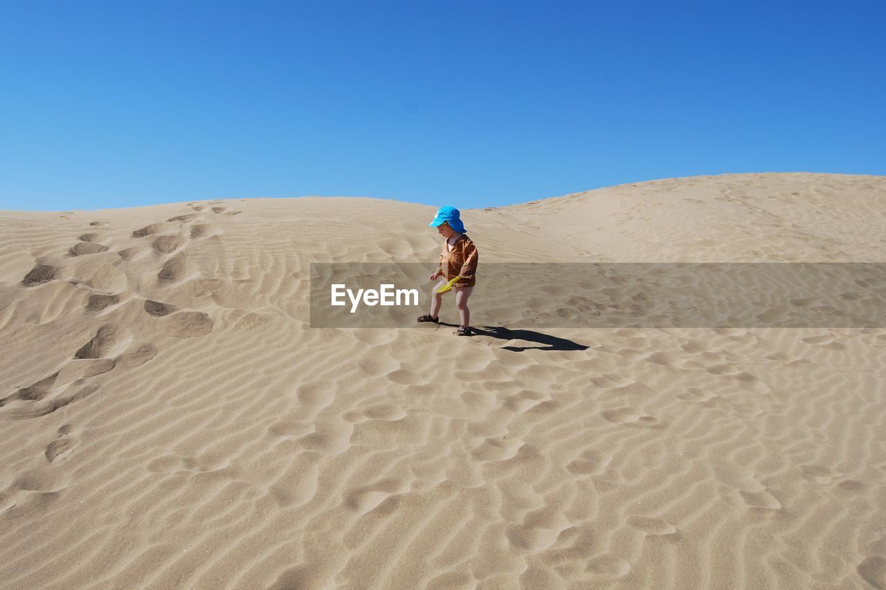 Boy standing on desert against clear blue sky during sunny day