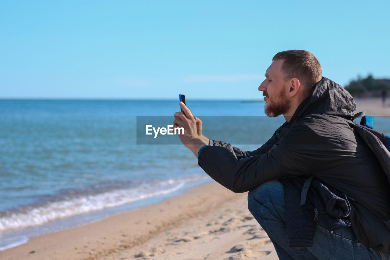 Bearded man in black raincoat on blue sea landscape background with smartphone in hands takes selfie