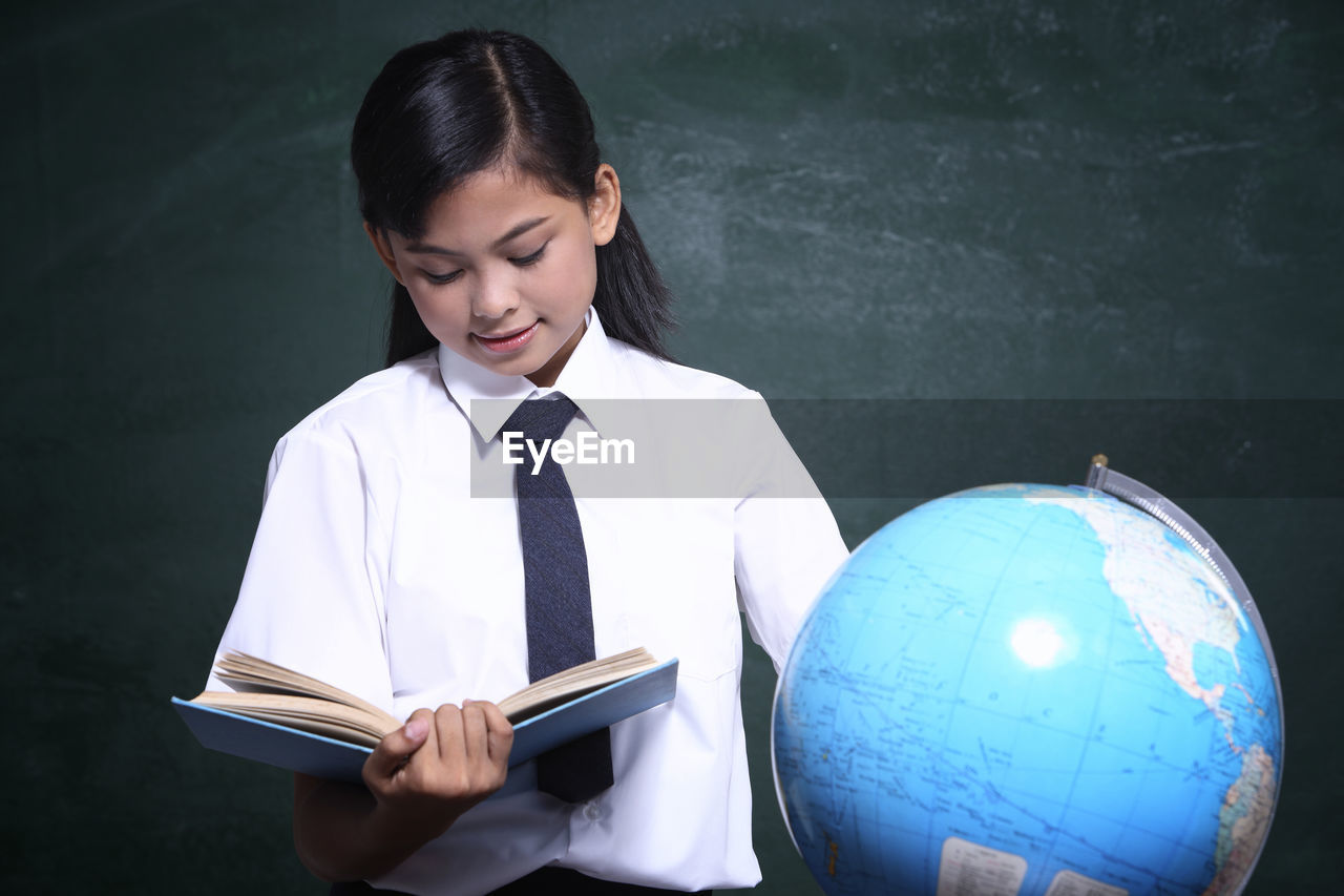 Schoolgirl reading book by globe against blackboard