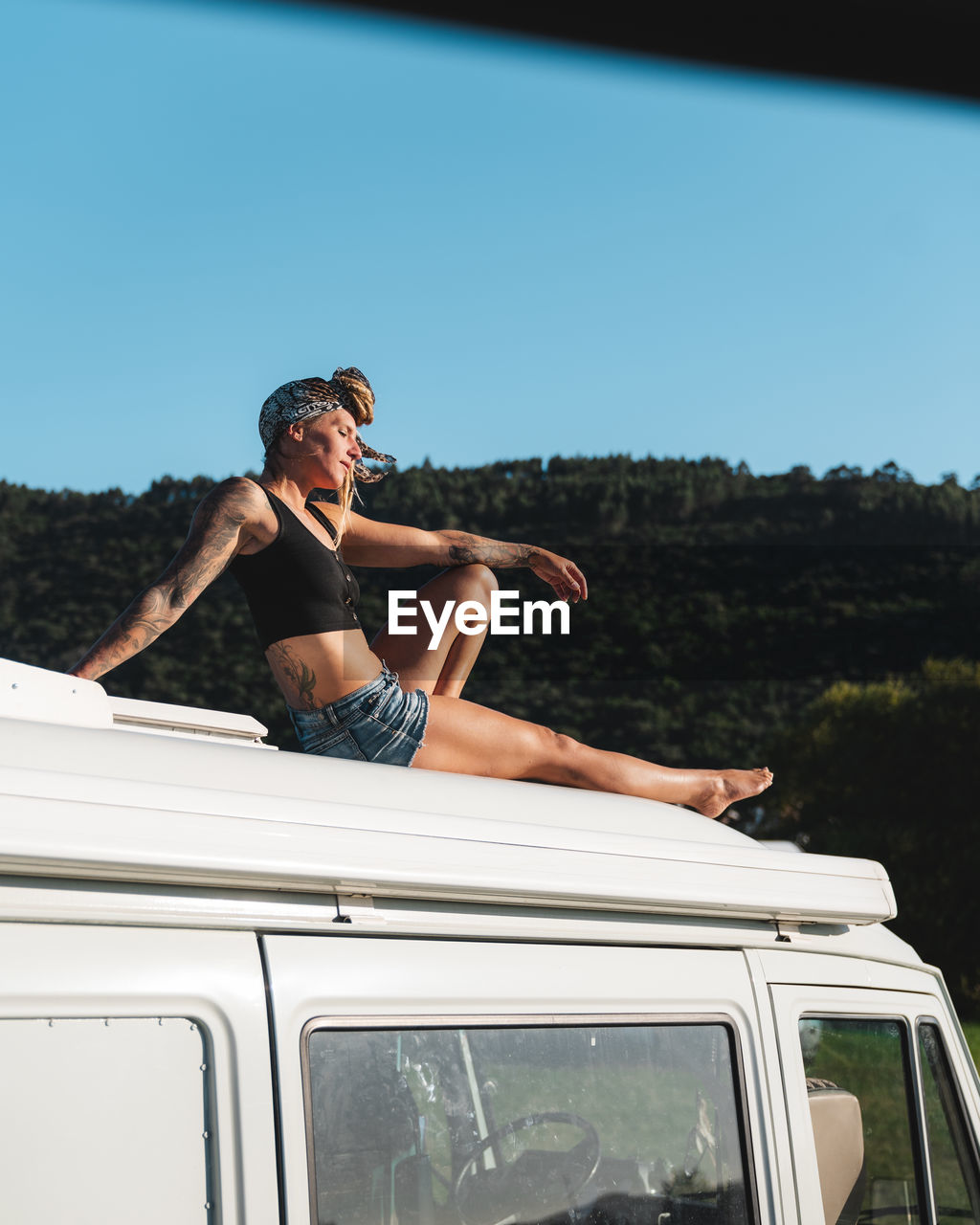 Full body side view of of pleasant barefoot thoughtful hippie female traveler sitting with eyes closed on roof of white camping van parked in nature during road trip