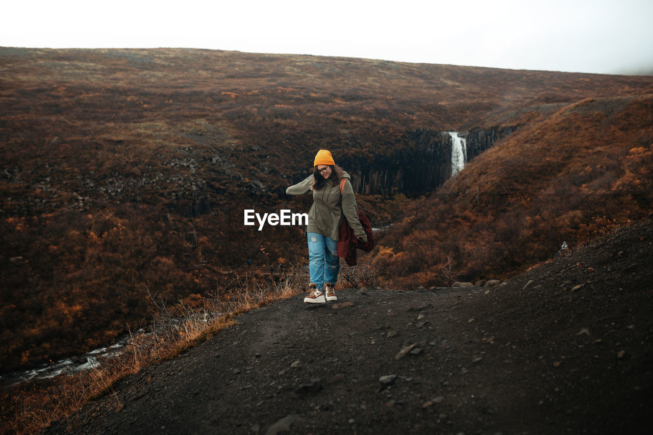 Young tourist in eyeglasses and hat with piercing looking down on hill near waterfall and mountain river