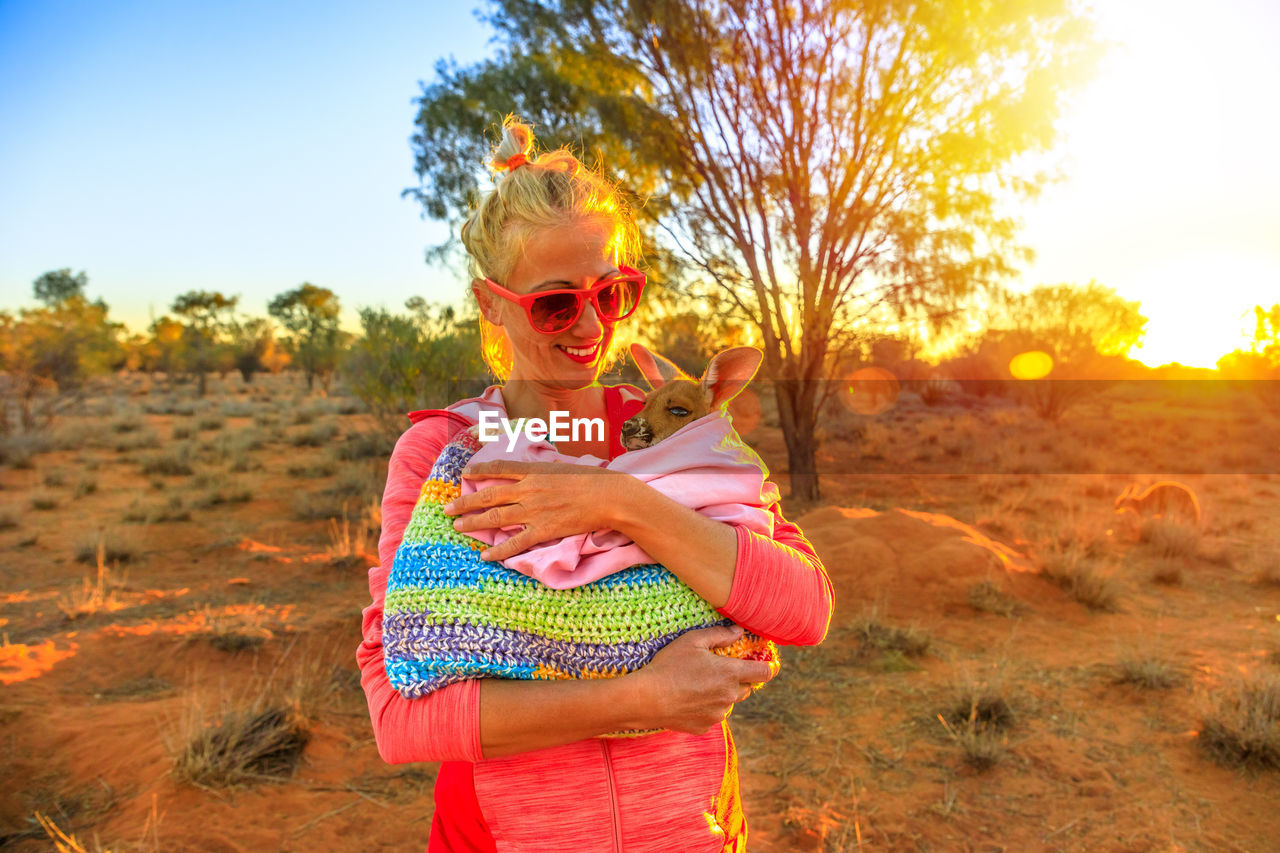 Smiling woman holding kangaroo on field during sunset