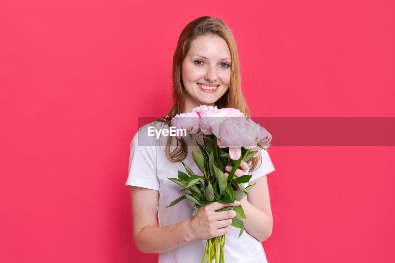 flower, pink, flowering plant, women, plant, smiling, bouquet, holding, one person, flower arrangement, looking at camera, portrait, happiness, adult, studio shot, emotion, colored background, waist up, indoors, female, rose, beauty in nature, young adult, positive emotion, freshness, nature, front view, bride, bunch of flowers, celebration, red, love, standing, cheerful, event, pink background, long hair, copy space, lifestyles, hairstyle, flower head, dress, person, valentine's day, brown hair, photo shoot