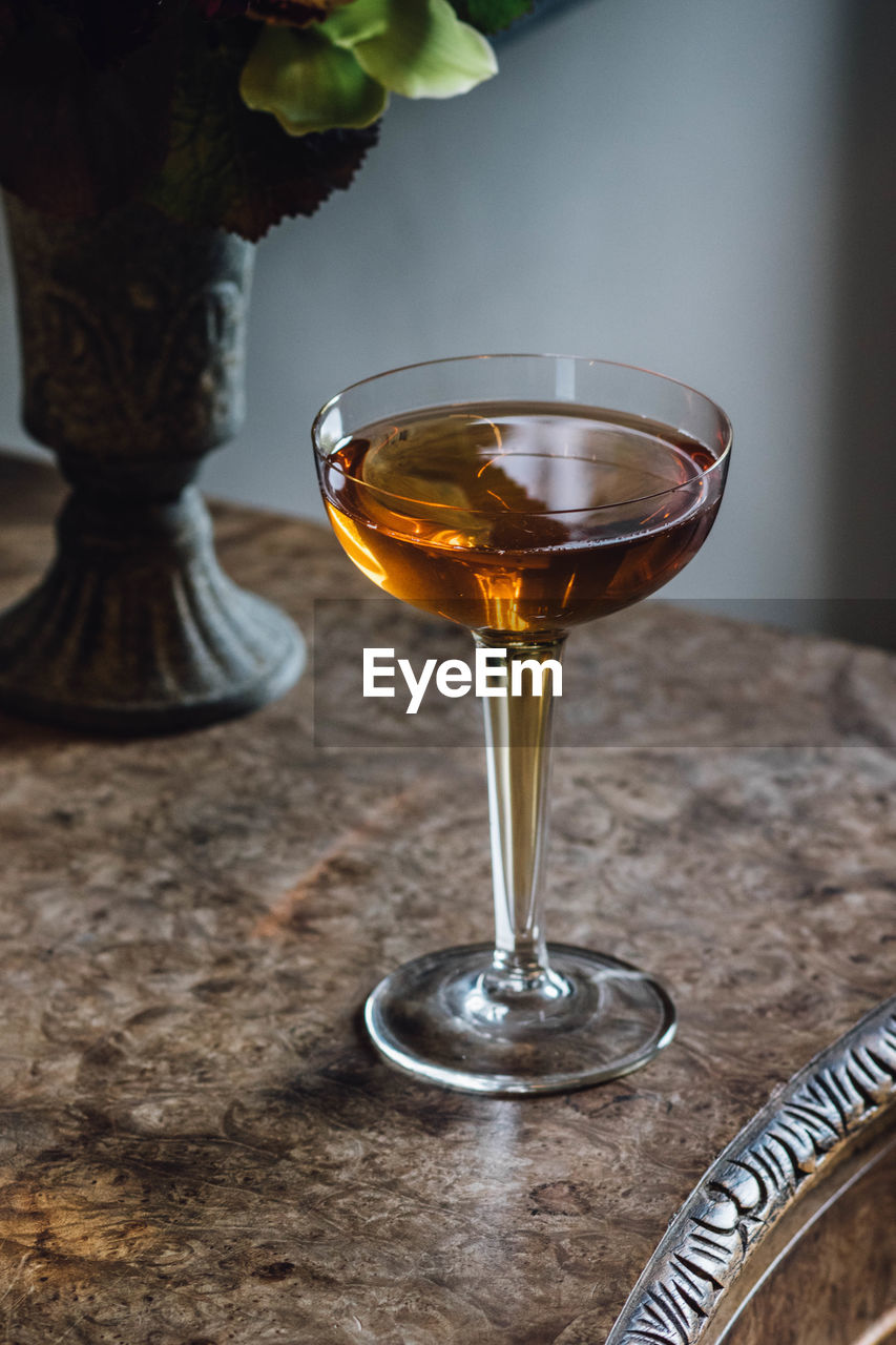 Illuminated whiskey drink in hollow stem coupe glass on burl wood table with bouquet in vase