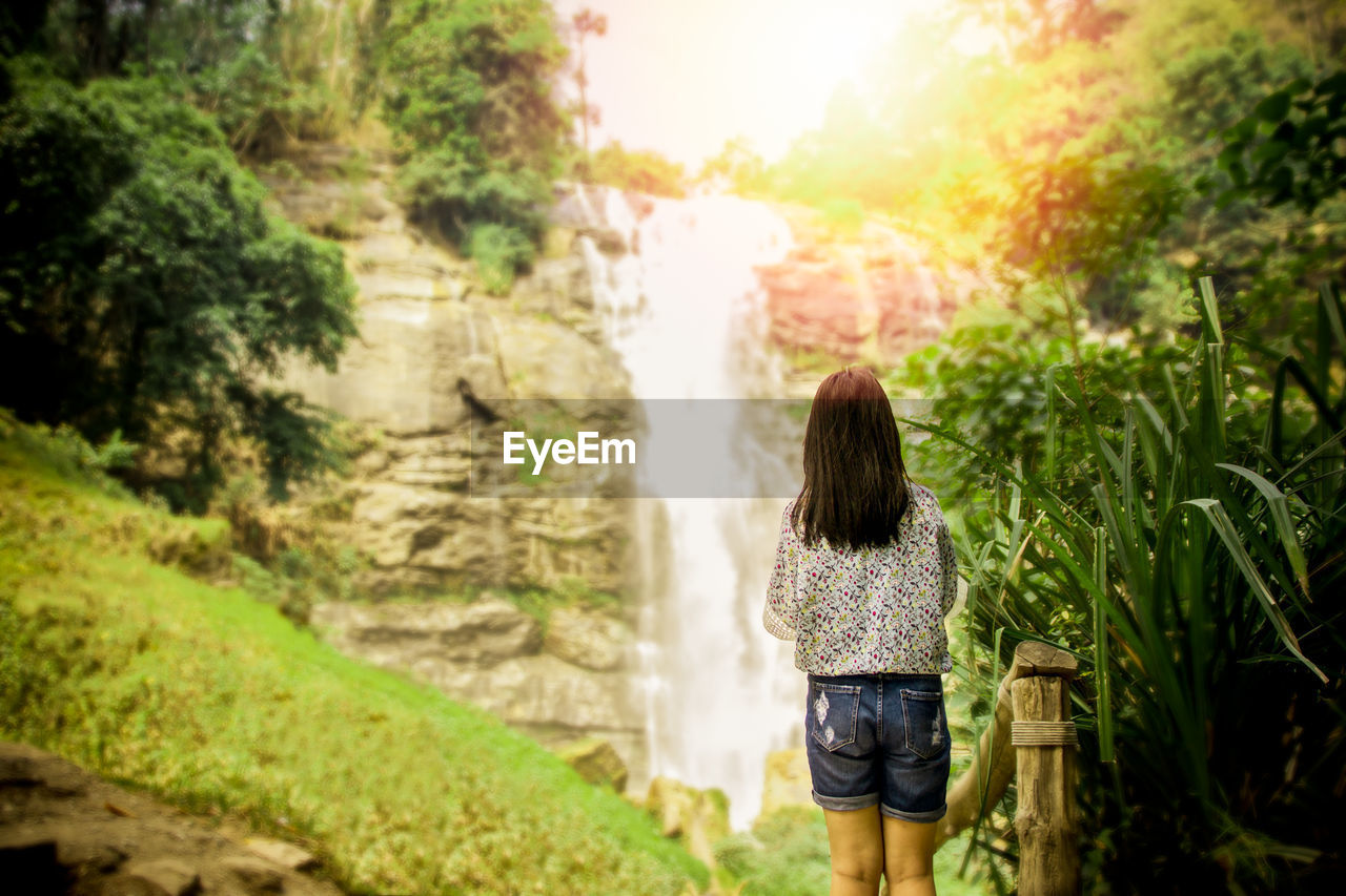 Rear view of teenage girl standing against waterfall