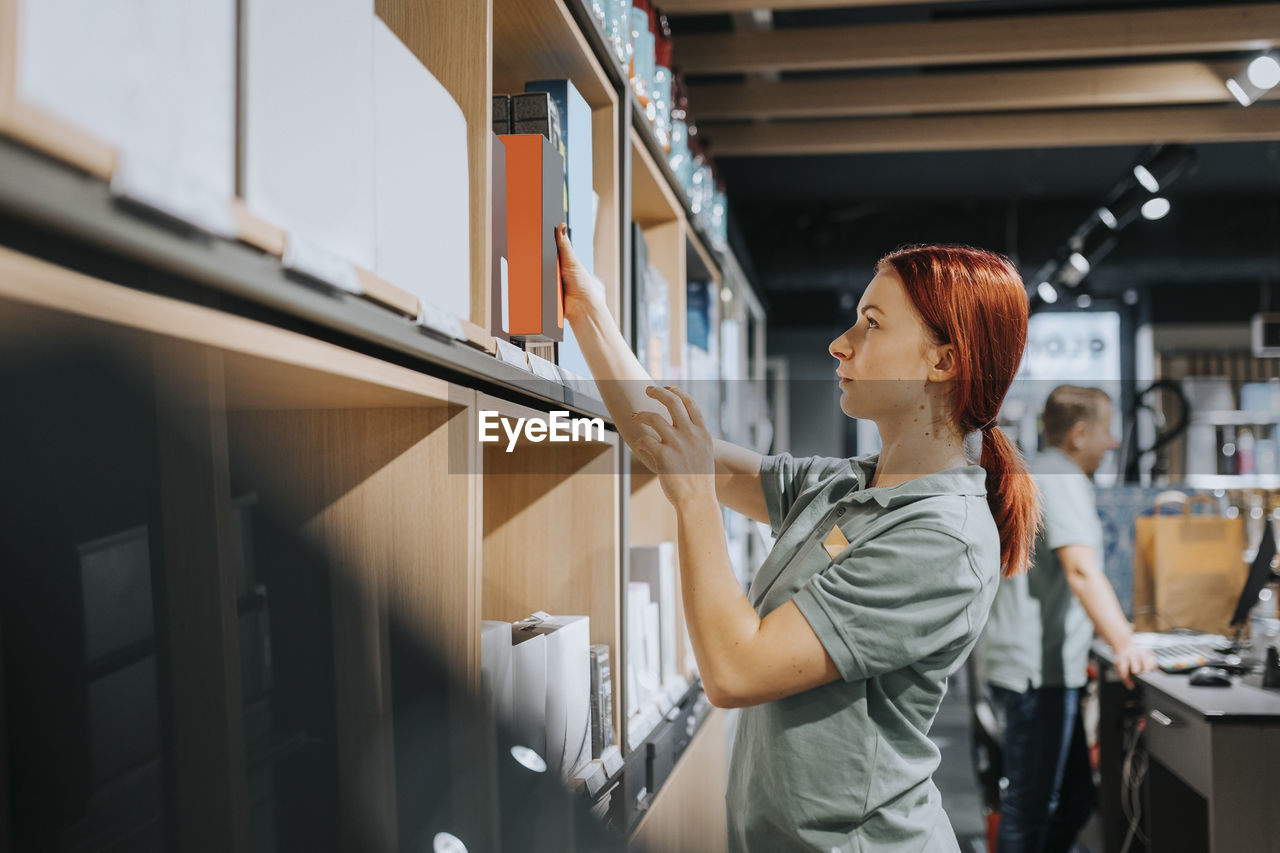 Side view of redhead saleswoman arranging appliance on shelf while working in electronics store