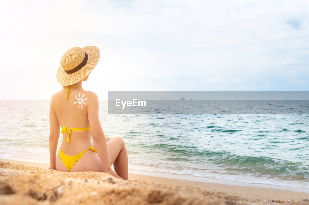 A young caucasian woman in a swimsuit and a straw hat sits on the beach in front of the sea with her