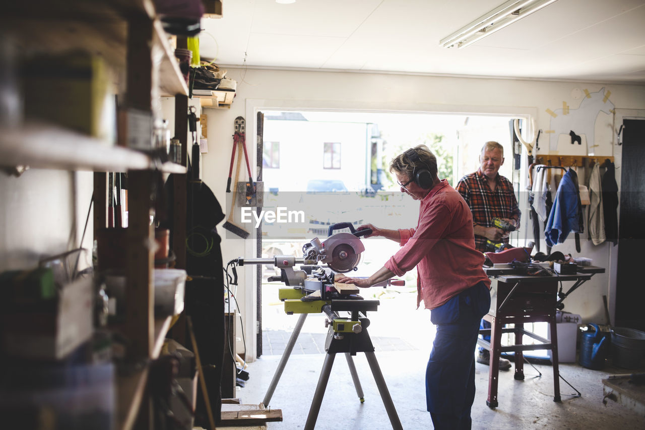 Side view of senior woman using circular saw while man holding drill in background