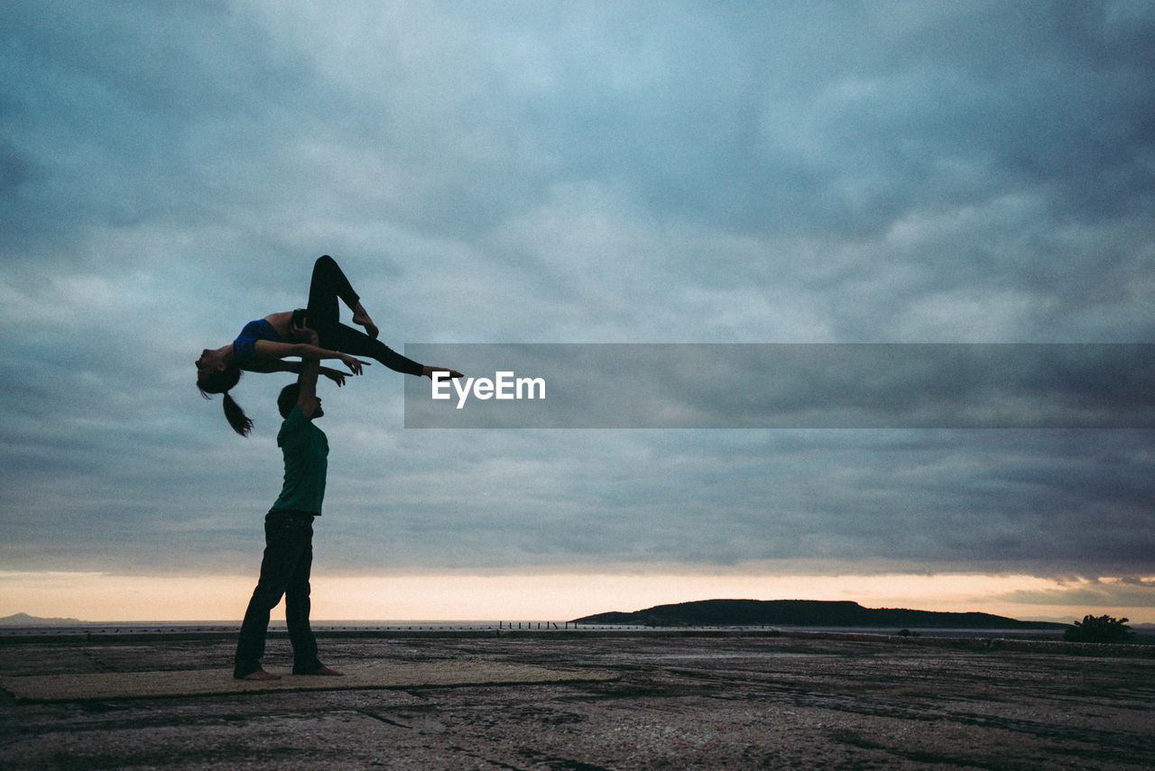 Full length of man balancing woman on hand by sea against cloudy sky