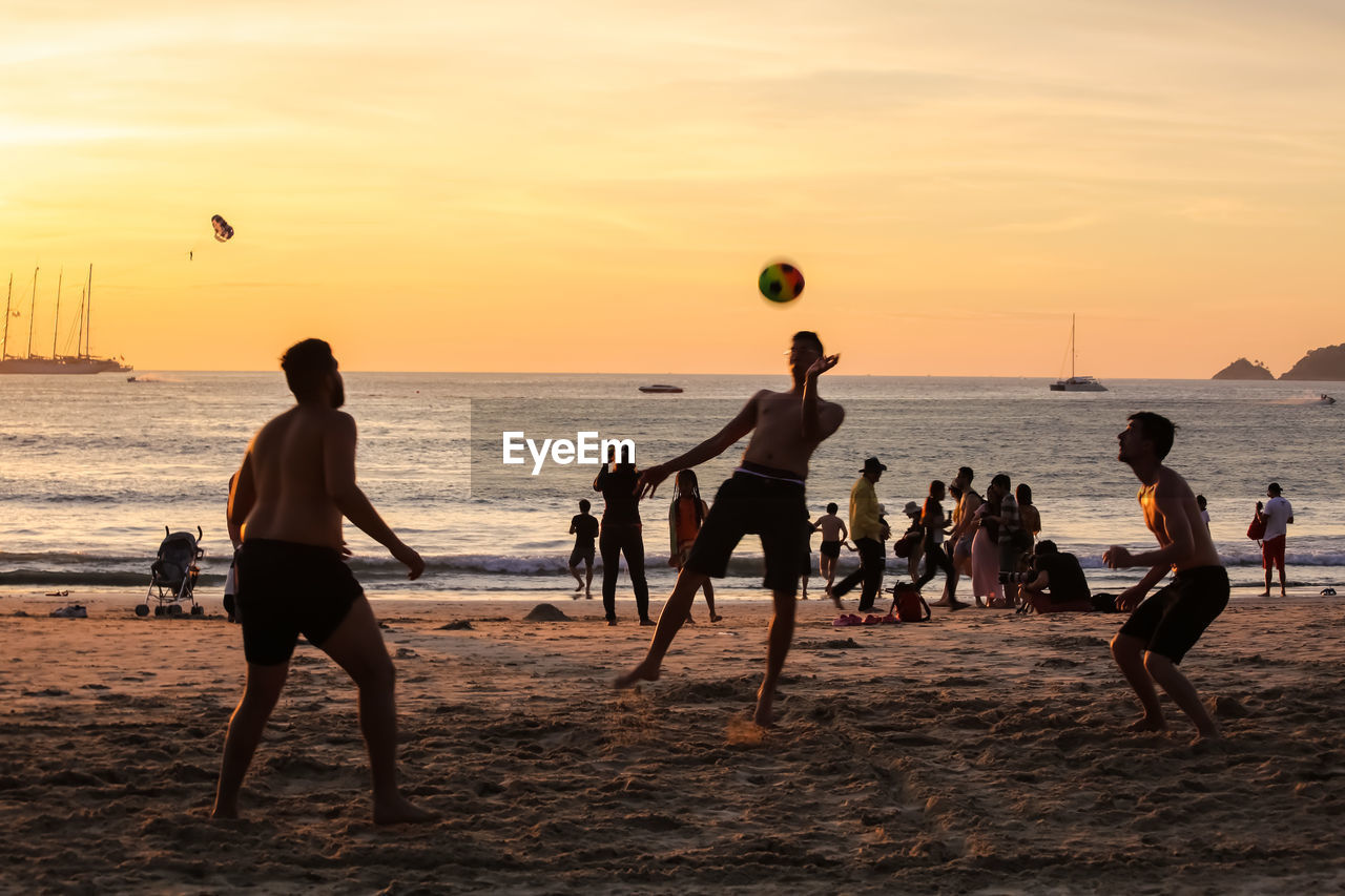 PEOPLE PLAYING WITH BALL ON BEACH AGAINST SUNSET SKY