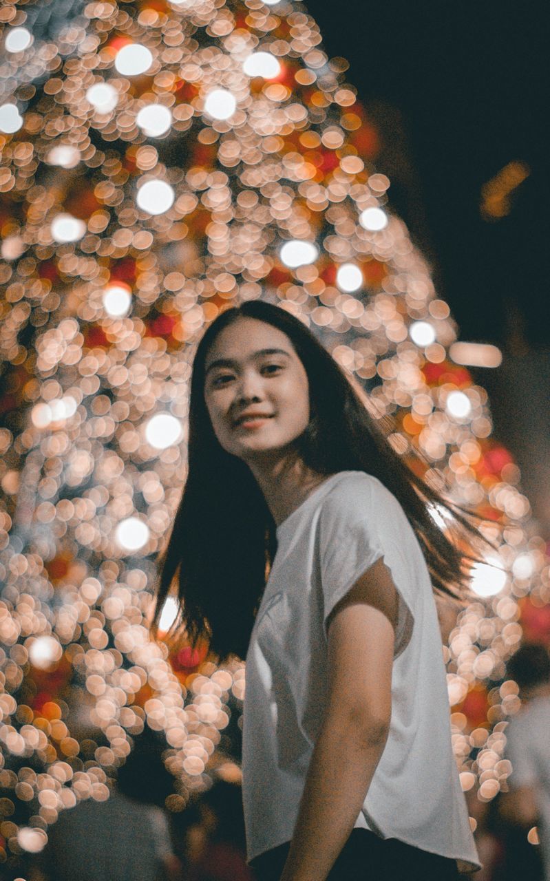 Portrait of young woman standing against illuminated christmas tree at night