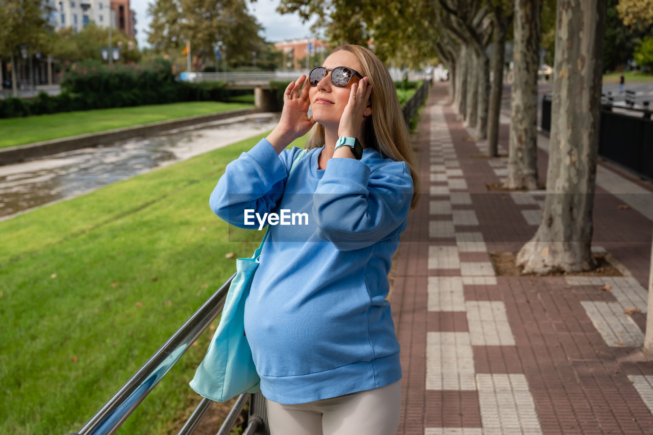 Pregnant woman looks at the sun in sunglasses. mom-to-be in a blue hoodie walks around the city
