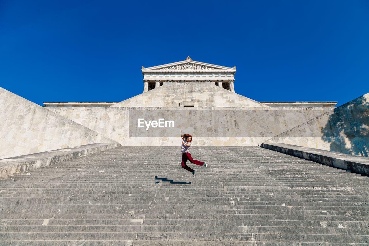 Low angle view of happy woman jumping on staircase against clear blue sky