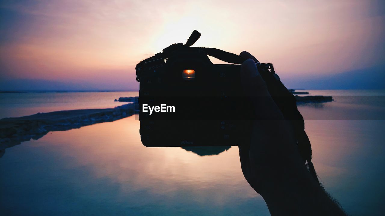 Cropped hand of man photographing sea against cloudy sky during sunset from camera