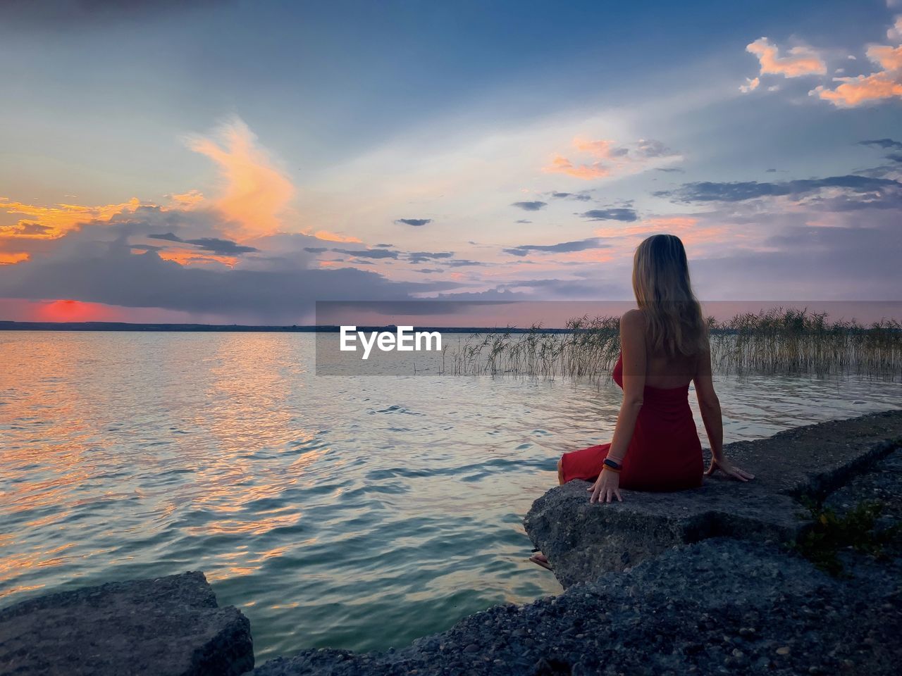 Rear view of woman in red dress sitting down near the lake at sunset and looking at the sky