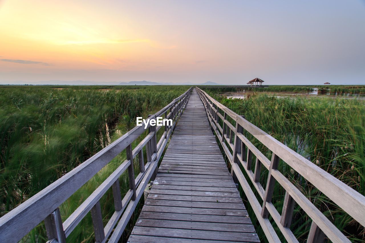 Wooden boardwalk amidst field against sky during sunset