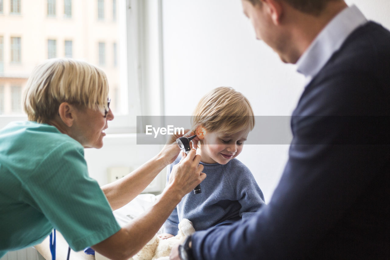 Female doctor examining boy's ear with otoscope while sitting by father in hospital