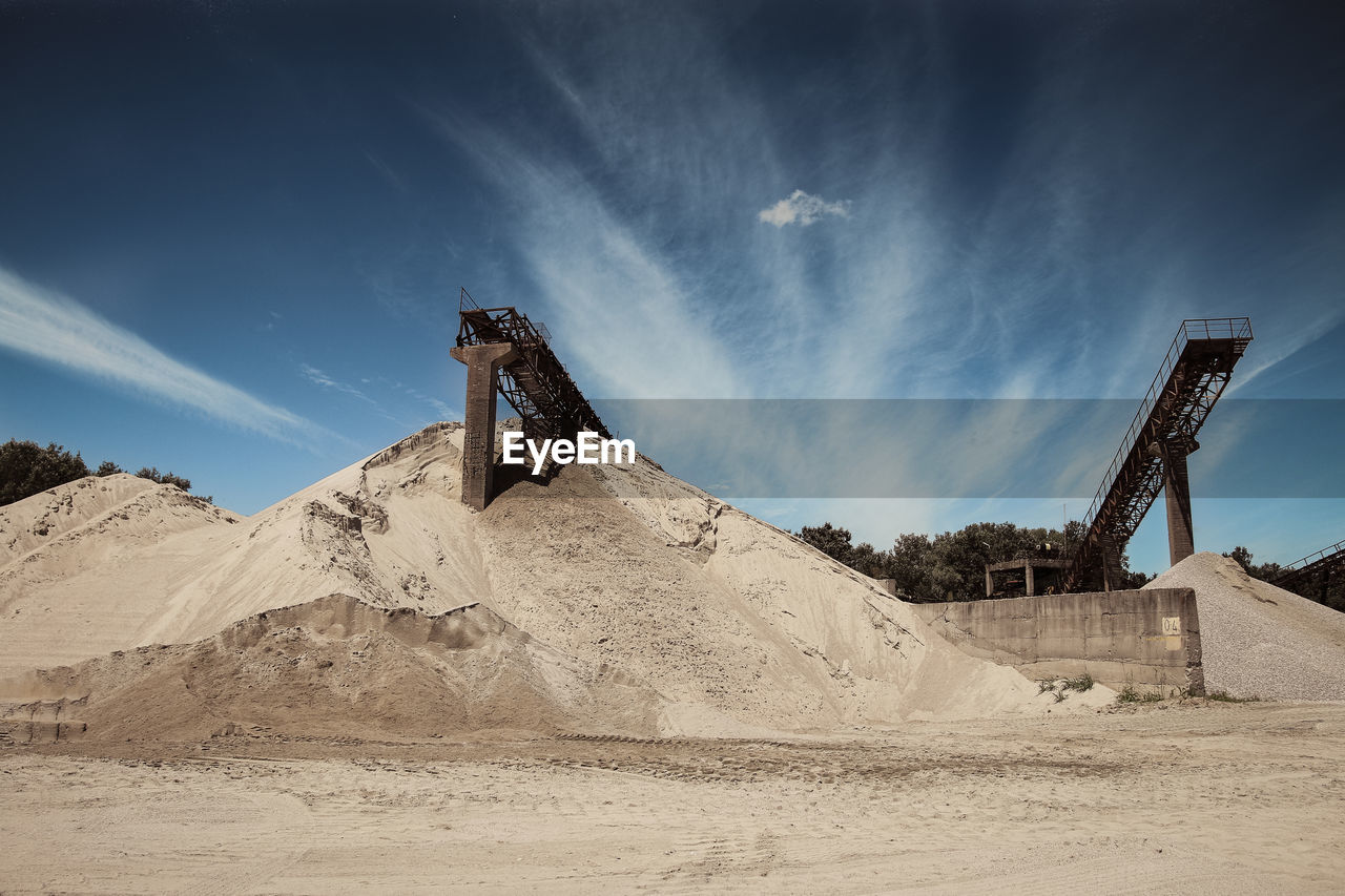 Low angle view of conveyor belt on sand pile at mine against sky