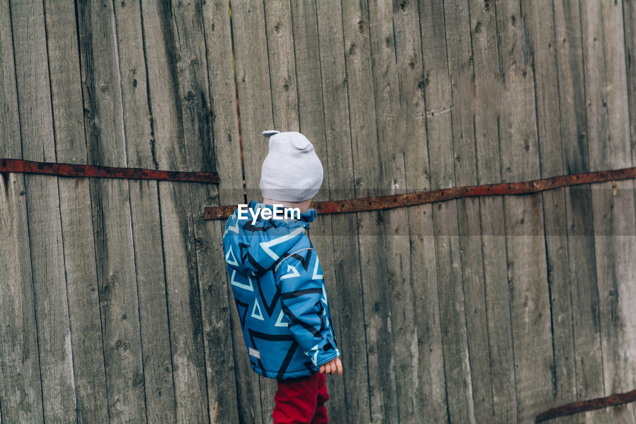 A child in a blue jacket and a hat walks near a wooden wall. real and serene people