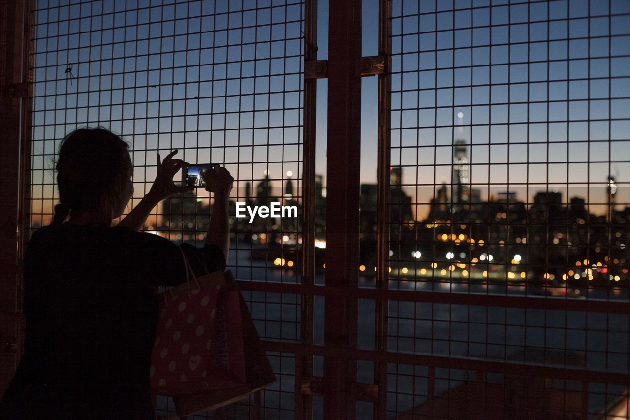 Silhouette woman photographing illuminated city from metal grate