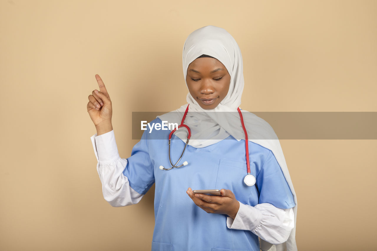 Online nurse with headscarf isolated on yellow background.holds smartphone, points finger up person