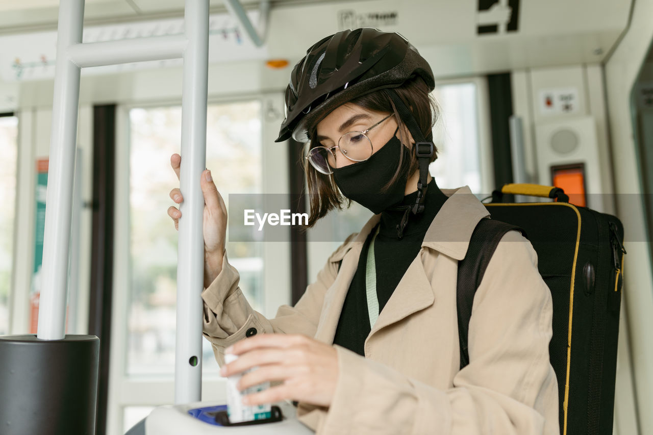 Woman wearing face mask and cycling helmet putting validating ticket in machine while inside tram