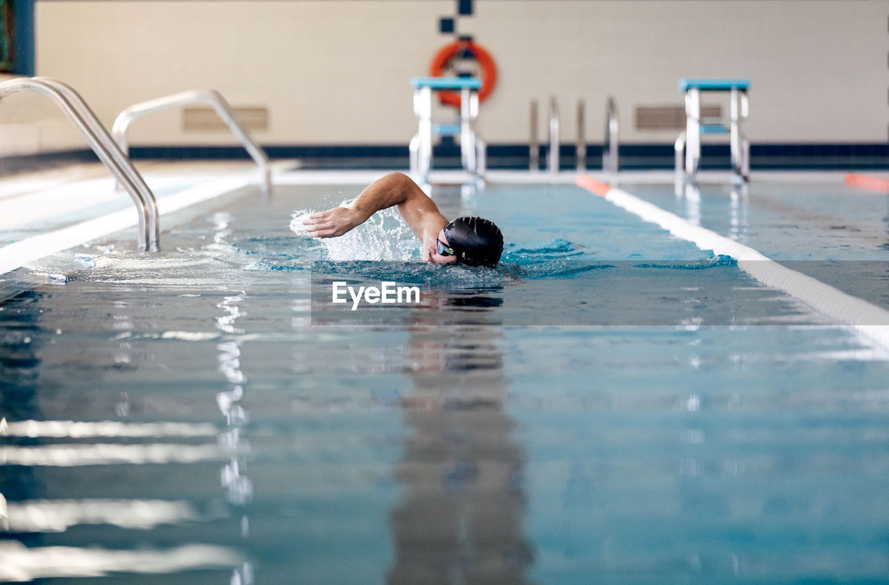 Male swimmer in cap and goggles performing sidestroke while swimming in rippled pool