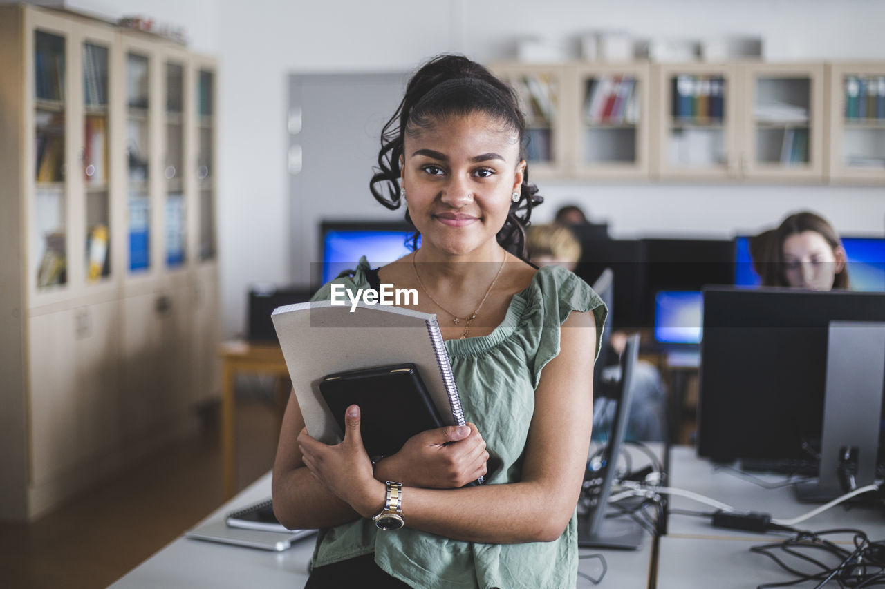 Portrait of smiling teenage girl standing with book in computer lab at high school