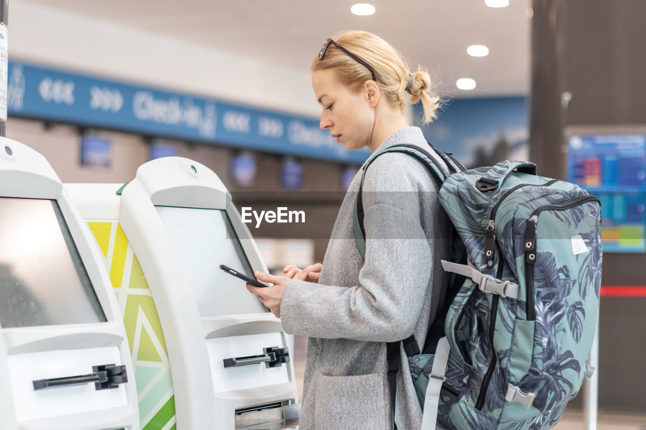 Smiling woman using smart phone while standing by atm machine