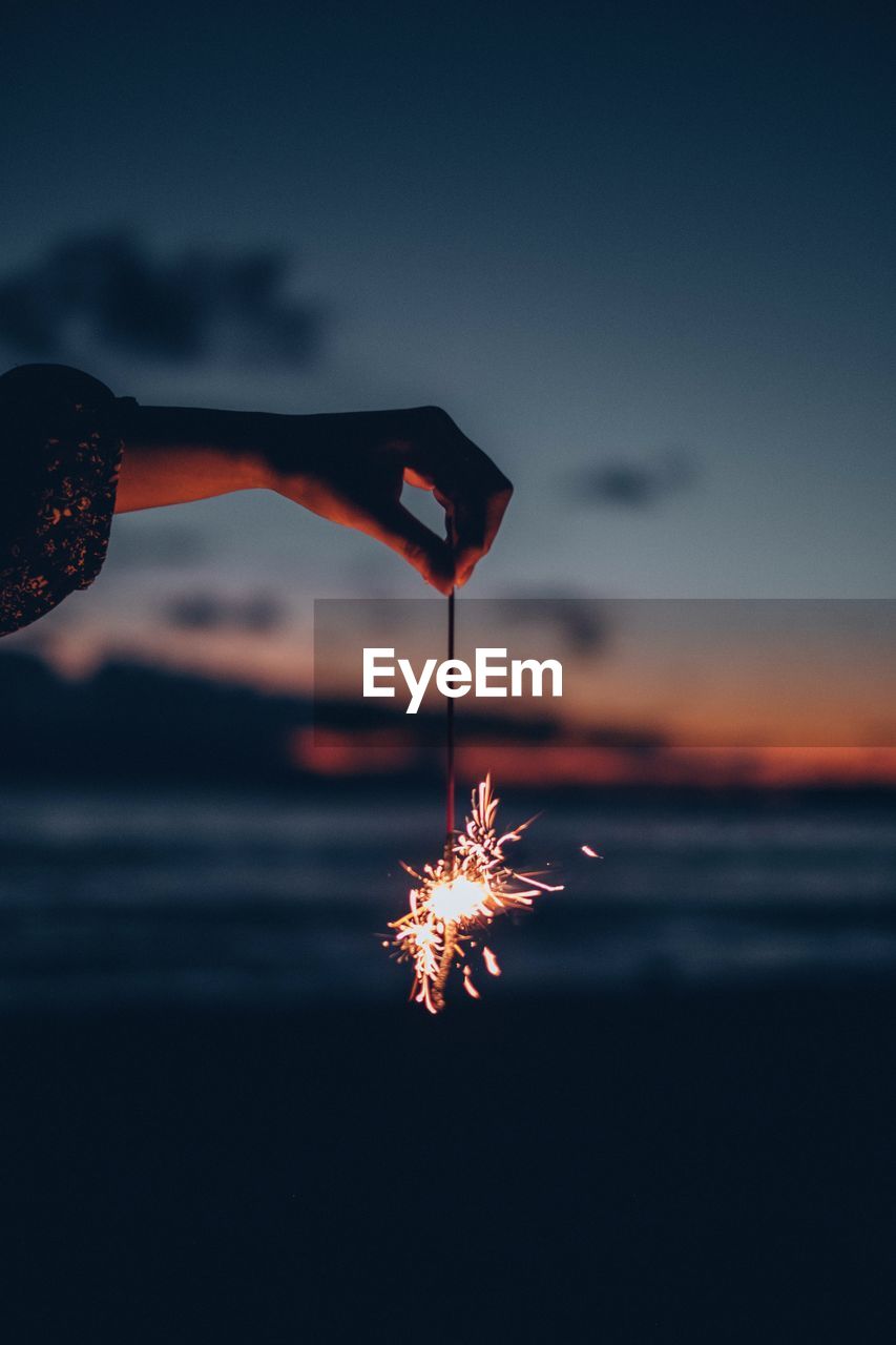 Cropped hand holding burning sparkler against sky during sunset