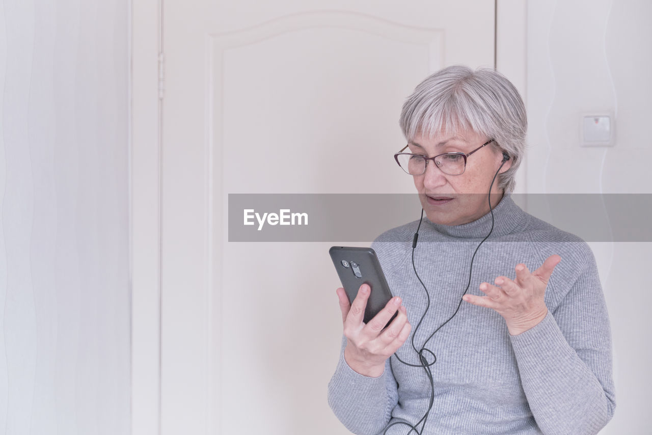 A gray-haired senior woman with glasses and a gray turtleneck talking on mobile phone at home. 