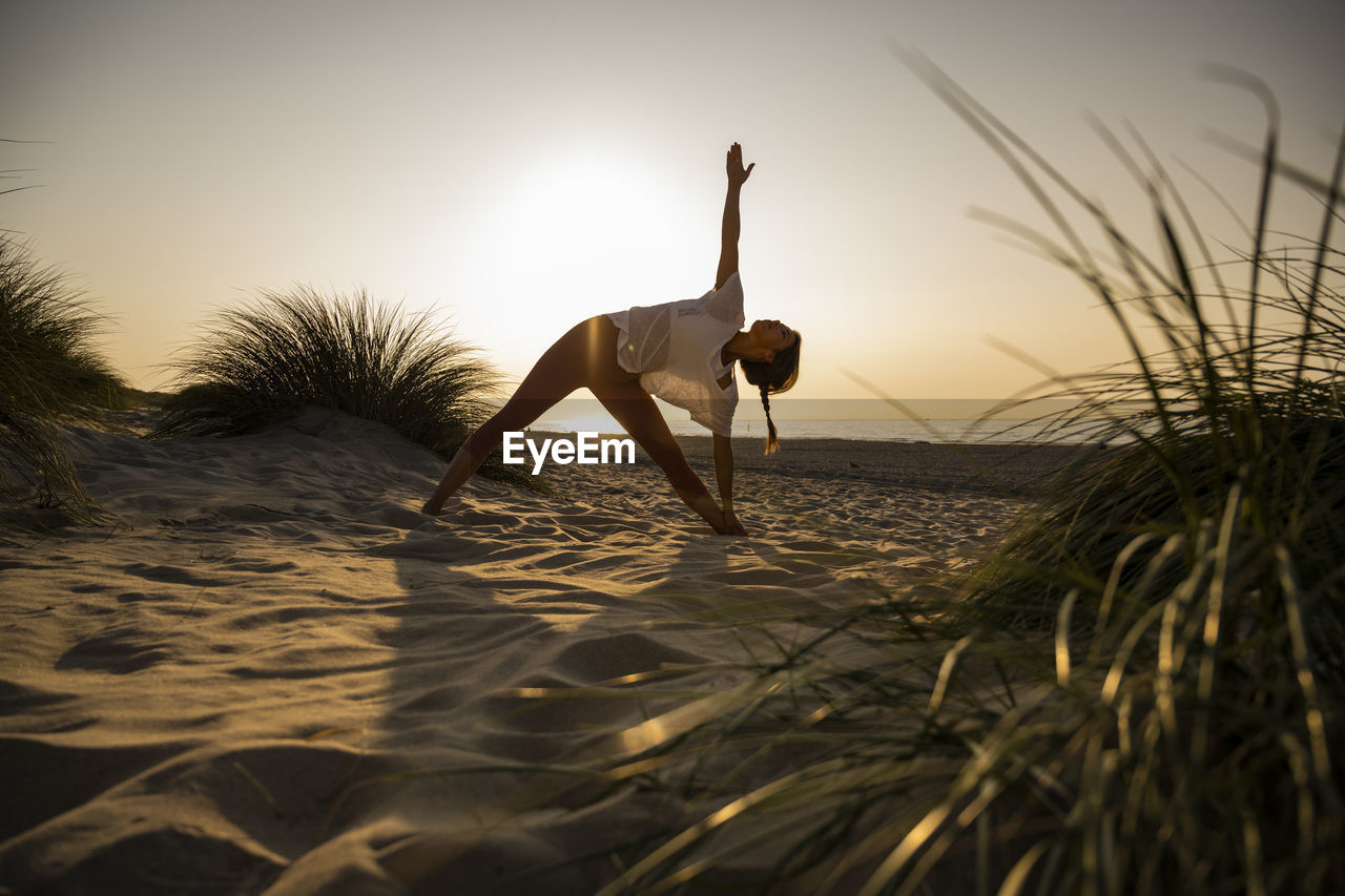 Young woman practicing triangle position yoga amidst plants at beach against clear sky during sunset