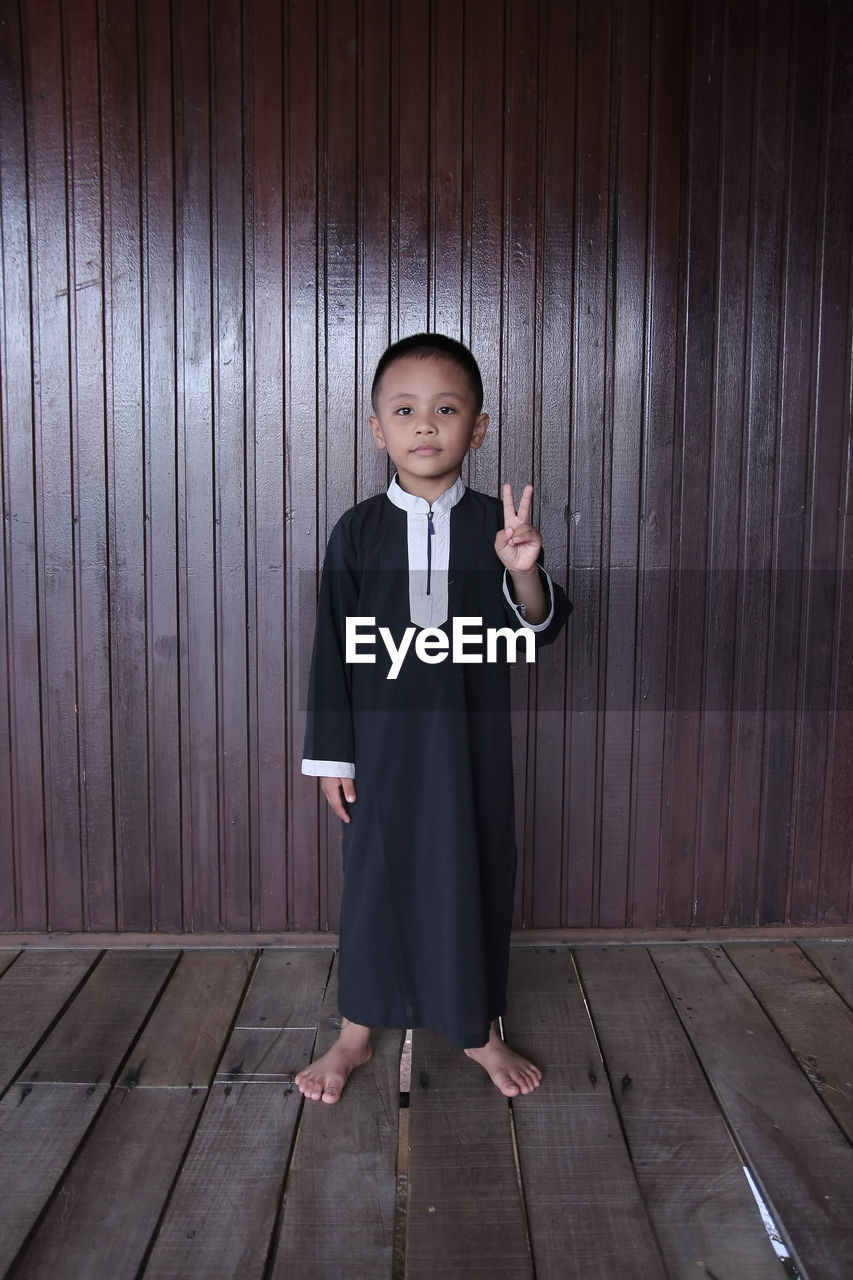 Portrait of boy showing peace sign while standing against wooden wall