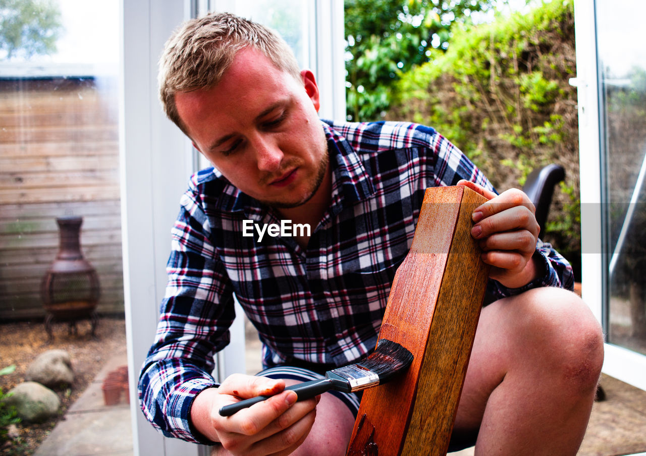 Young man painting wooden plank while crouching in yard