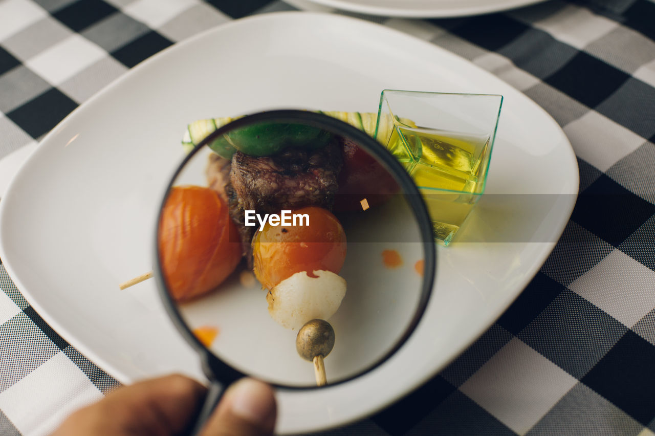 Close-up of cropped hand holding magnifying glass on food served in plate