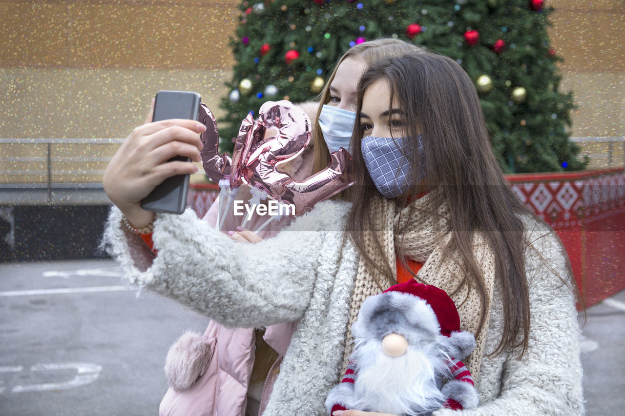 Women wearing mask photographing with mobile phone against christmas tree in winter
