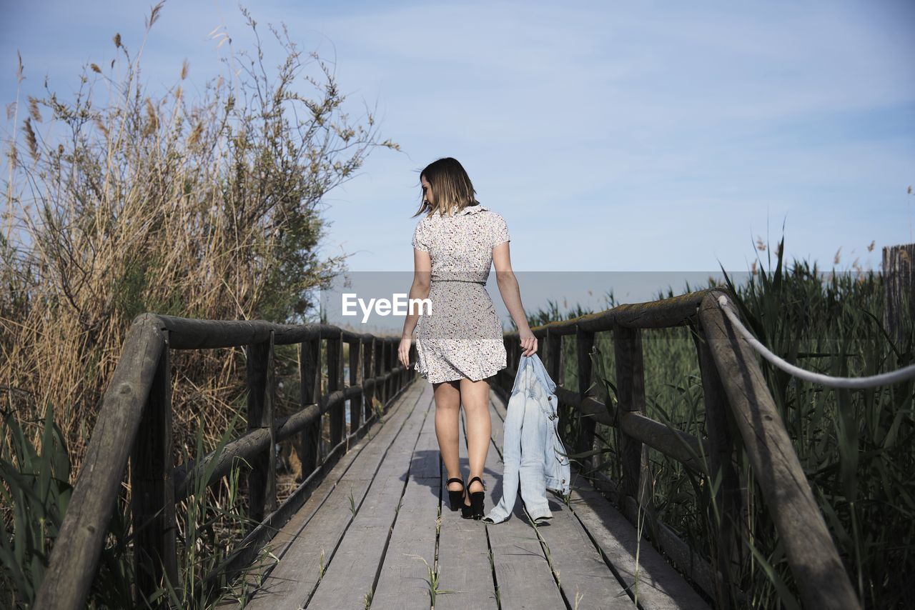 Full body back view of young female in casual dress with jacket in hand strolling on wooden footbridge among tall dry grass in sunny day