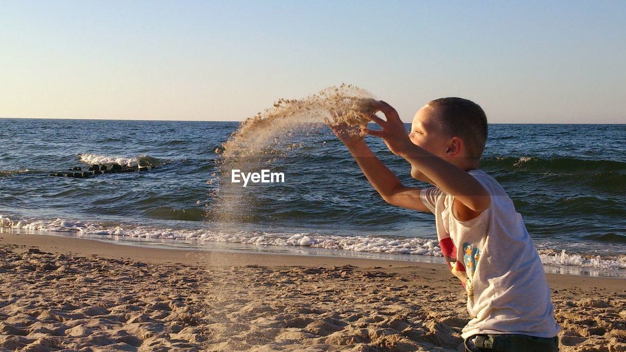 Boy throwing sand at beach against clear sky