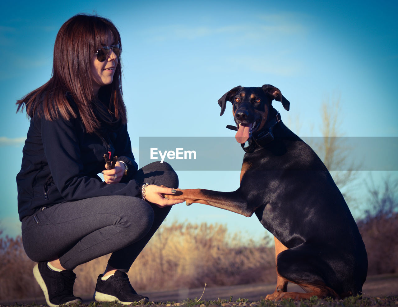 PORTRAIT OF WOMAN WITH DOG SITTING AGAINST SKY