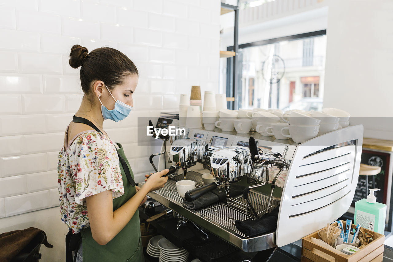 Young woman with face mask making coffee while standing in kitchen at coffee shop